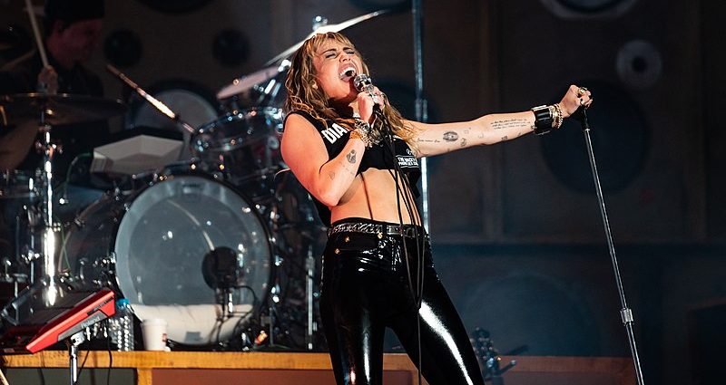 Miley Cyrus on stage holding a microphone, mouth open. She is wearing black shiny jeans and a black crop top