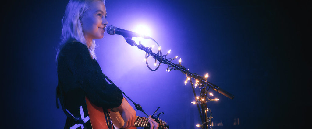 Phoebe Bridgers singing into a microphone whilst playing the guitar