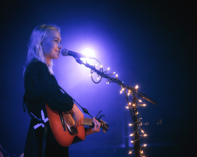 Phoebe Bridgers singing into a microphone whilst playing the guitar
