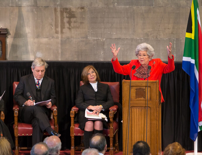 Baroness Betty Boothroyd wearing a red suit, standing behind a podium with arms raised.