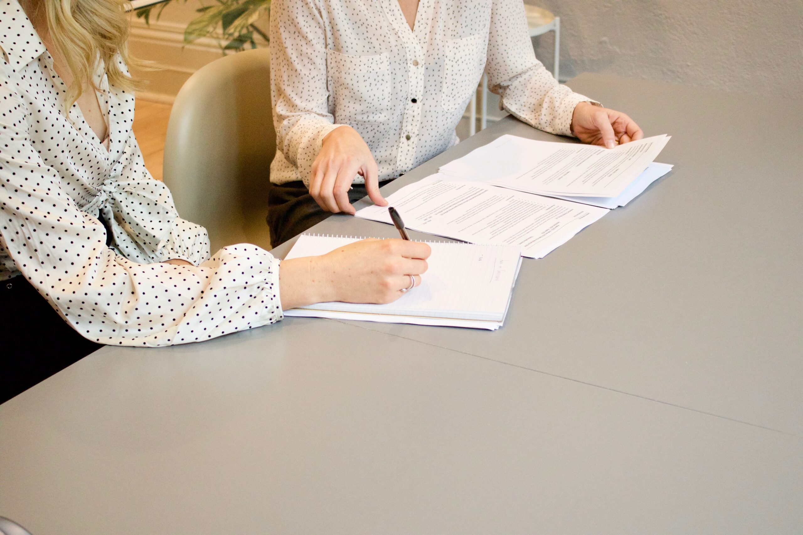 Two women signing papers at desk. The image is used to show women in a business setting, as happens in The Apprentice.