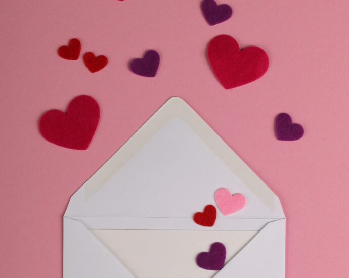 An open white envelope laid flat against a pink background. Emerging from the envelope are some carefully-cut pink and purple paper hearts. The image is used to reflect the love and emotion that Sabrina Carpenter put into her album, based on emails she sent to herself.
