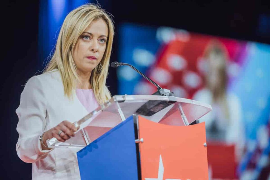 Image of the Prime Minister of Italy, Giorgia Meloni, standing behind a podium at a conference. The blonde haired woman is wearing a white long sleeved dress.