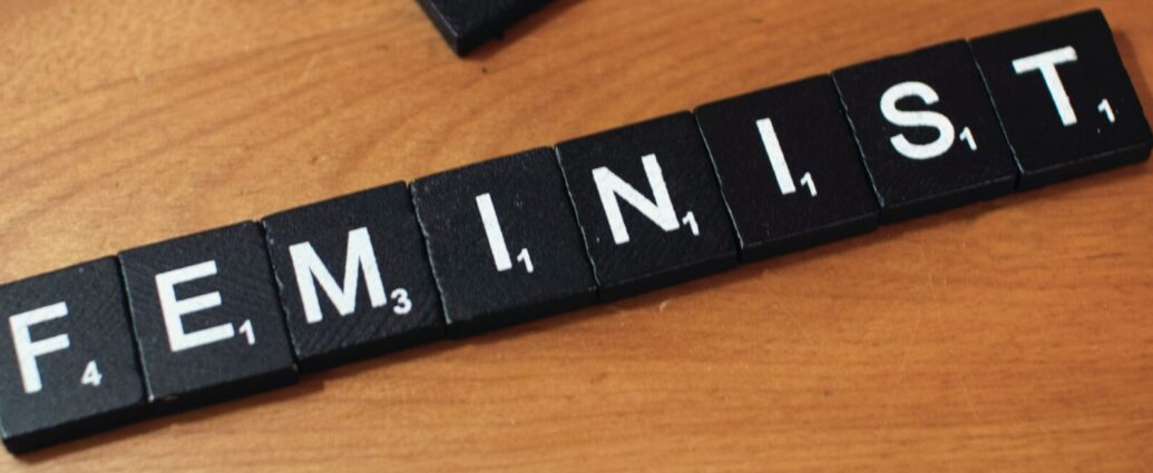 The word 'feminist' spelled out using black and white Scrabble tiles on a wooden surface.