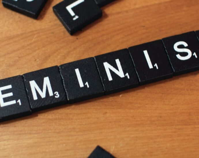 The word 'feminist' spelled out using black and white Scrabble tiles on a wooden surface.