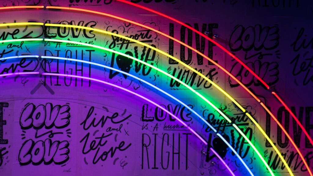 A neon pride rainbow against a darker black and purple background featuring slogans including 'love wins'. this represents the messages in songs by LGBTQ+ artists.
