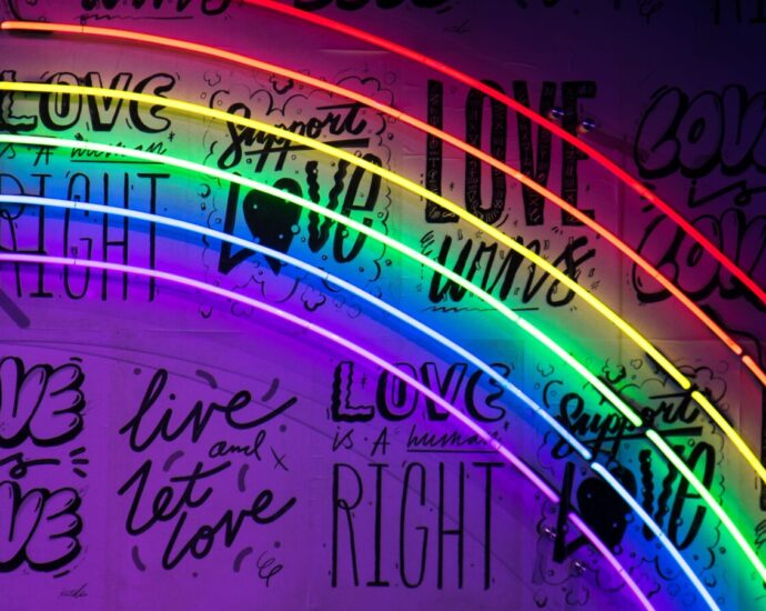 A neon pride rainbow against a darker black and purple background featuring slogans including 'love wins'. this represents the messages in songs by LGBTQ+ artists.