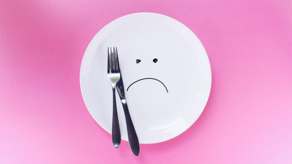 A plate with a sad face drawn on it, with a knife and fork, on a pink background.