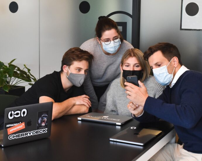 Four people looking at a phone screen. They all wear a mask.