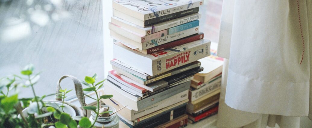 A stack of books tidily placed on a white windowsill. There's a green plant on the left and a white curtain on the right.