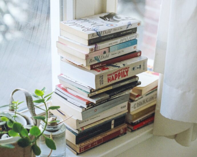 A stack of books tidily placed on a white windowsill. There's a green plant on the left and a white curtain on the right.
