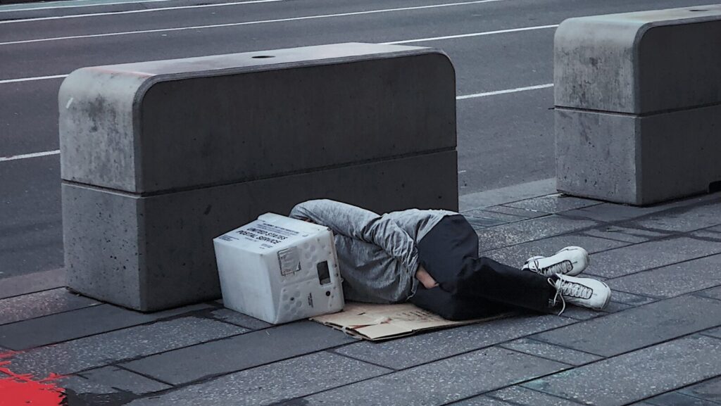 This image shows someone lying down on the floor next to what could be described as hostile architecture, because the seating area is small and has no back rest.