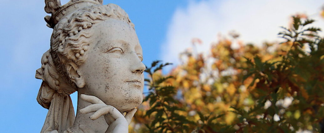 A stone, female statue stands under a blue sky, a hand placed thoughtfully under her chin.