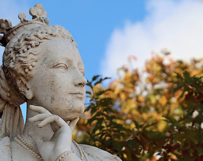 A stone, female statue stands under a blue sky, a hand placed thoughtfully under her chin.