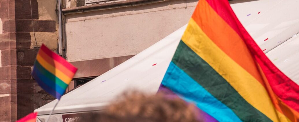 A rainbow pride flag is waving above a crowd of people. Flags like these can be seen in Heartstopper season two
