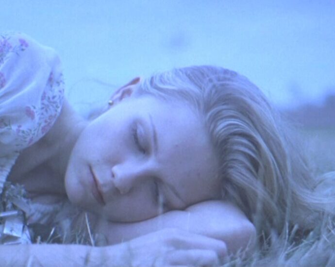 A close up of a blond girl's face as she lies in some grass with her eyes shut.