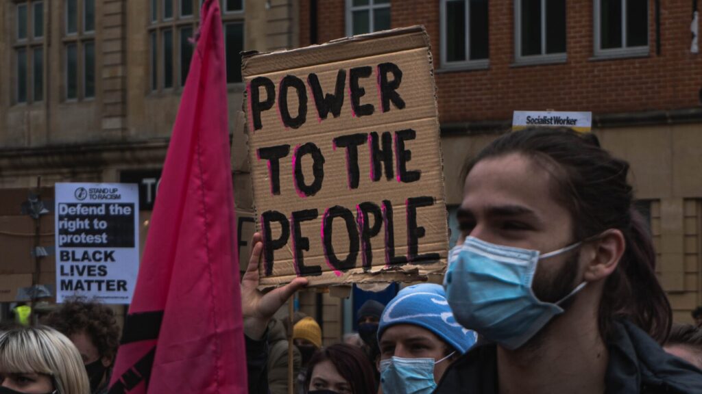 Image shows protestors at a Black Lives Matter protest holding a pink flag and two signs that read "POWER TO THE PEOPLE" and "Defend the right to protest. BLACK LIVES MATTER"