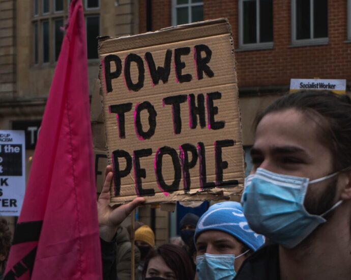 Image shows protestors at a Black Lives Matter protest holding a pink flag and two signs that read "POWER TO THE PEOPLE" and "Defend the right to protest. BLACK LIVES MATTER"