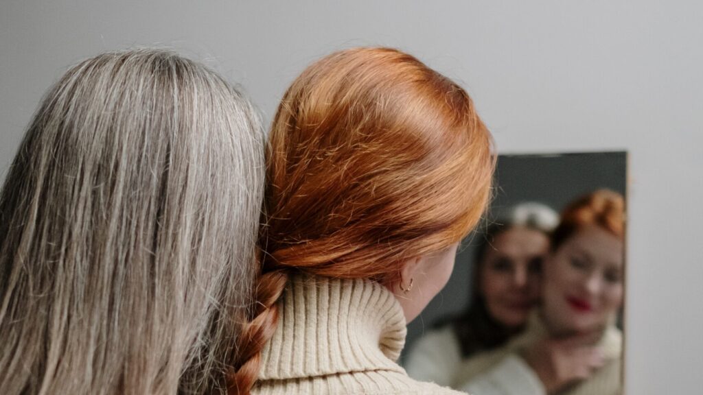Two White women, stand side-by-side looking at a mirror. One has gray hair, the other is a redhead. The photo is more focused on the back of their heads showcasing their different hair.