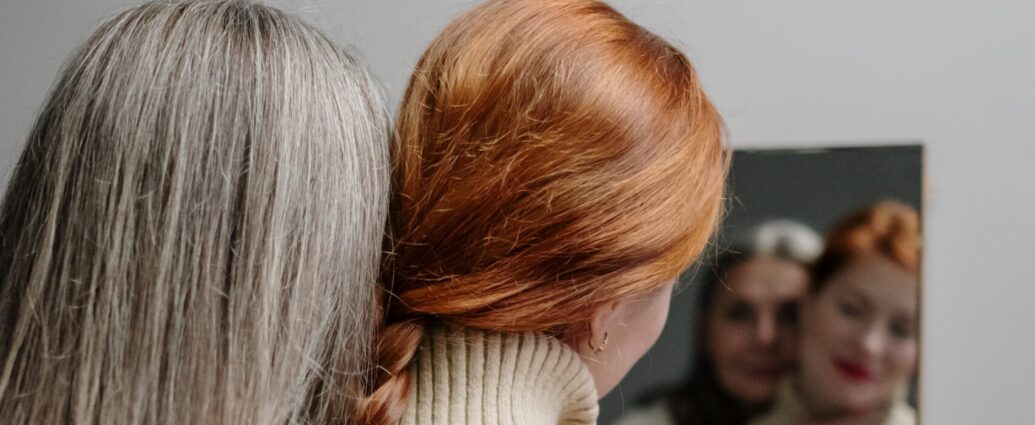Two White women, stand side-by-side looking at a mirror. One has gray hair, the other is a redhead. The photo is more focused on the back of their heads showcasing their different hair.