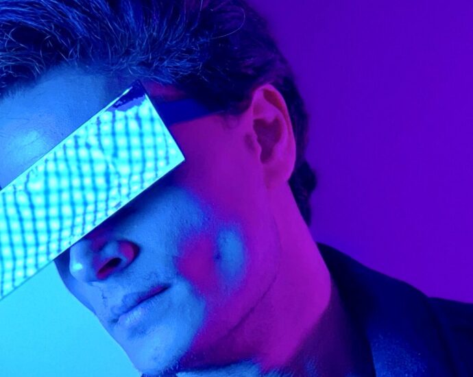 A man in a jacket wears some shades in a purple room.