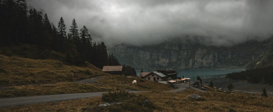 An eerie small town set into a mountainous area surrounded by trees and dark clouds, with a lake in the background.