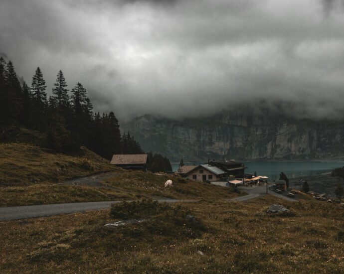 An eerie small town set into a mountainous area surrounded by trees and dark clouds, with a lake in the background.