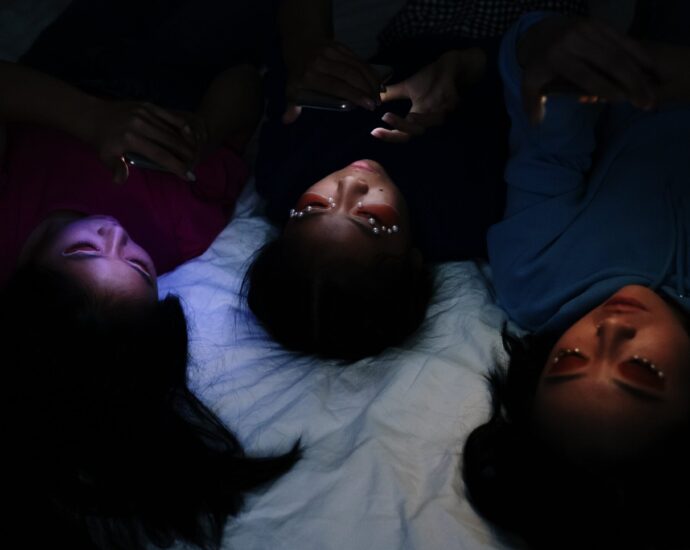 Image shows three women in ethereal 'Euphoria-esque' makeup lying on a bed, their faces lit by their phone screens.