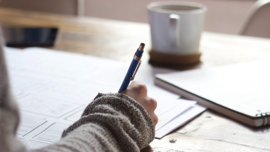 Person writing on brown wooden table near white ceramic mug. My journalism journey.