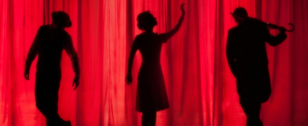 Three silhouettes standing in front of a red stage curtain.