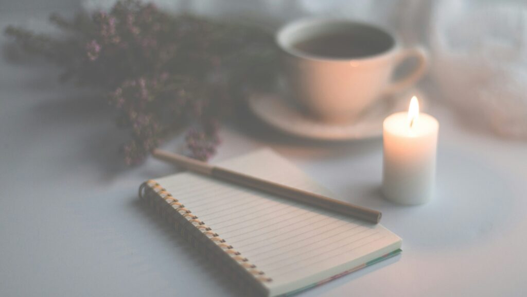 Image shows an open journal and pen, surrounded by a lit candle, cup of tea and dried flowers.