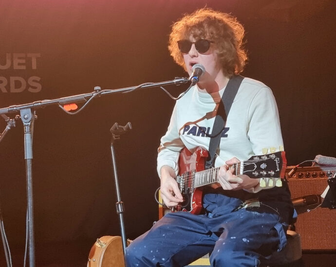 The Snuts member sat on stage wearing a white t-shirt, navy trousers and black sun glasses playing guitar and singing into a microphone.