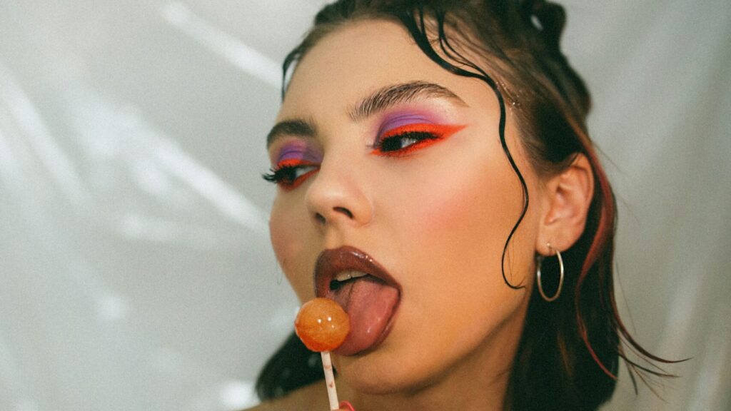 A woman with Y2K style waved hair, brown lipstick and brightly coloured eye makeup licks a lollypop.