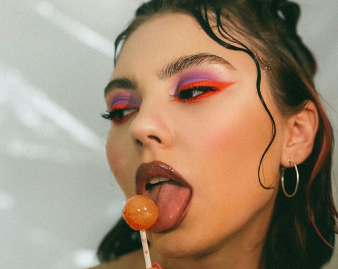 A woman with Y2K style waved hair, brown lipstick and brightly coloured eye makeup licks a lollypop.
