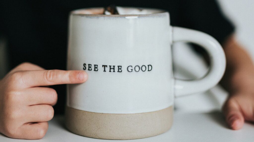 A finger is seen pointing at the words "see the good" engraved on a white mug.