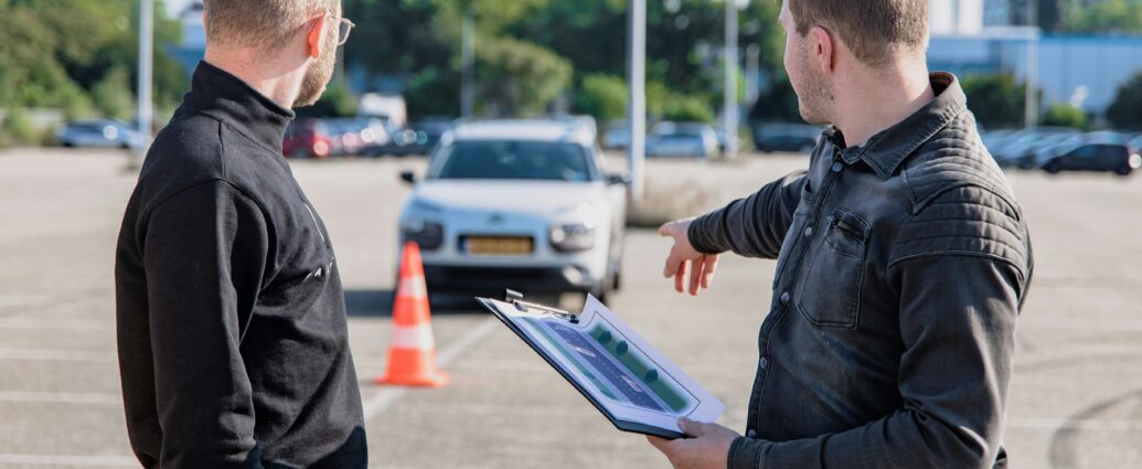 Driving instructor in a car park pointing to to a traffic cone with student and car in the background.