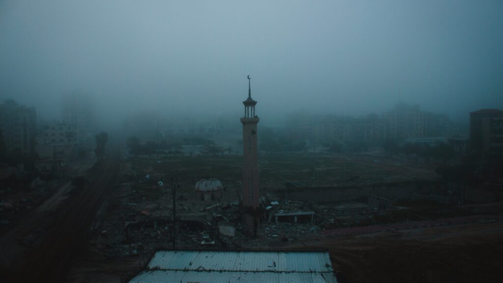 A photograph of a destructed part of the Gaza strip. A tower is seen as the only thing standing amongst rubble with a grey fog in the background.