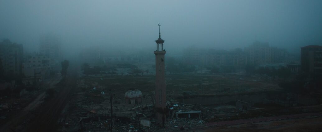 A photograph of a destructed part of the Gaza strip. A tower is seen as the only thing standing amongst rubble with a grey fog in the background.