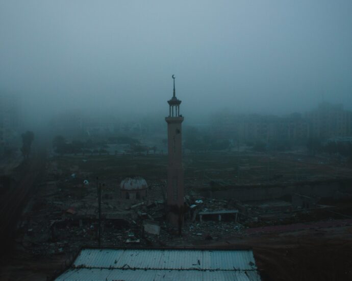 A photograph of a destructed part of the Gaza strip. A tower is seen as the only thing standing amongst rubble with a grey fog in the background.