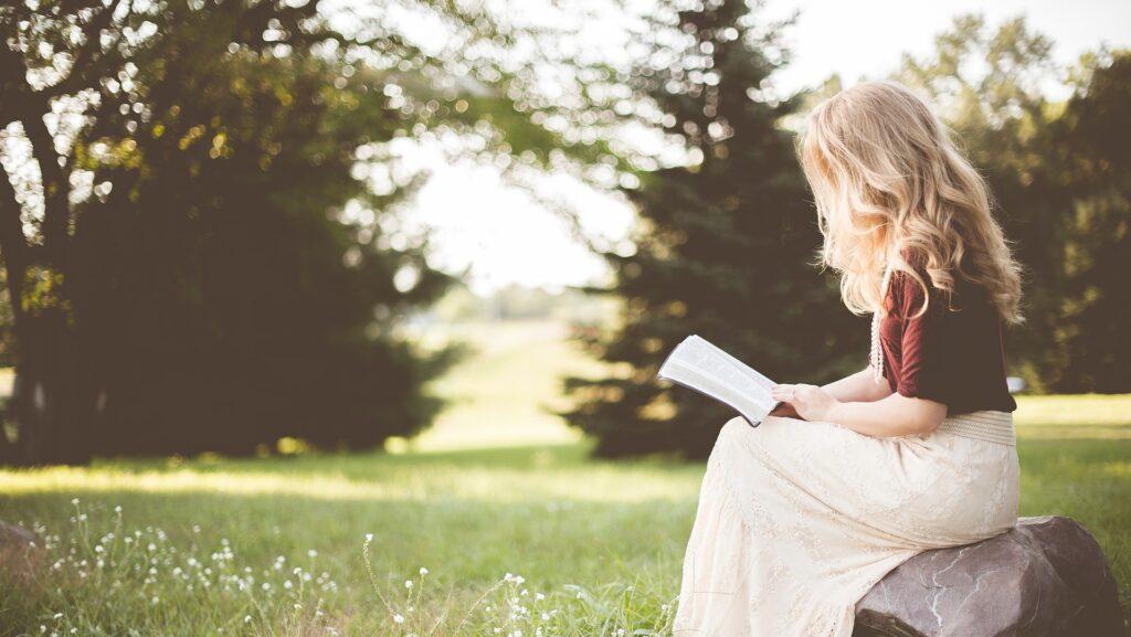 Girl sitting reading a book outside with grass and trees in the background potentially sunrise on the reaping