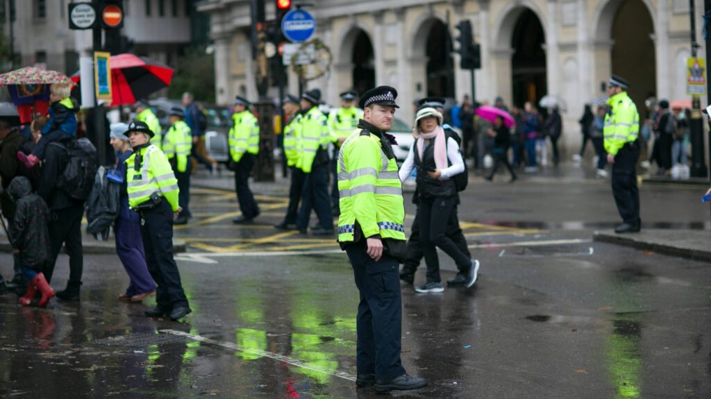 Police stand dispersed on a rainy street in London with a few members of the public walking around them to monitor child exploitation.