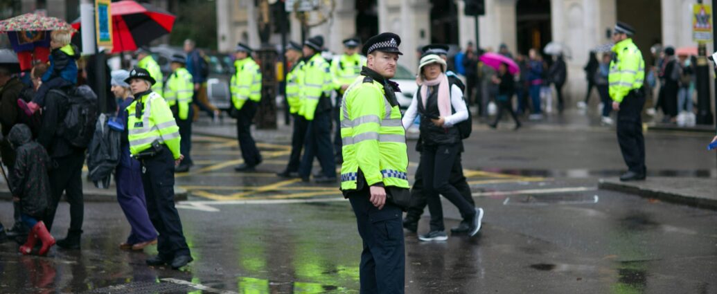 Police stand dispersed on a rainy street in London with a few members of the public walking around them to monitor child exploitation.