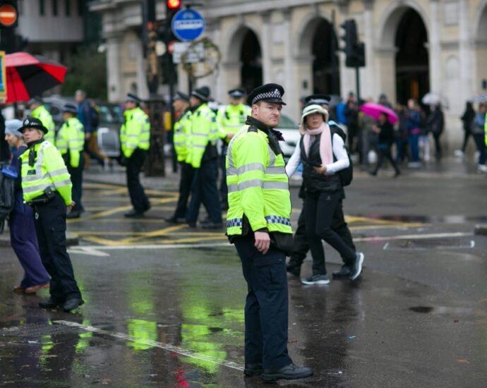 Police stand dispersed on a rainy street in London with a few members of the public walking around them to monitor child exploitation.
